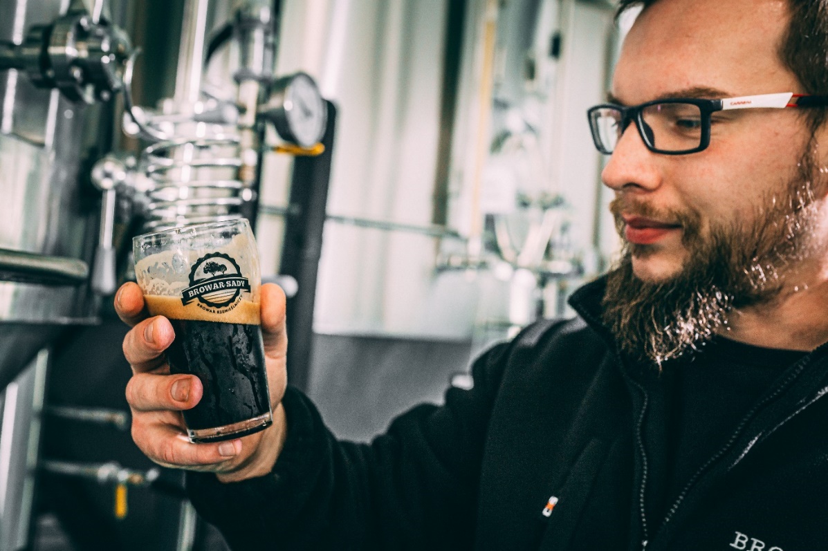 Maciej Wentowski trzymający w ręce małą szkalnkę z ciemnym piwem, w tle tanki z browaru Sady