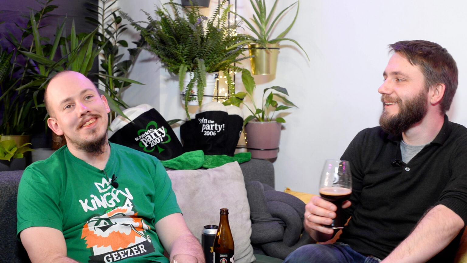 Grzegorz Stachurski (po prawej) i Łukas Szymański (po lewej) siedzą z głupimi minami na kanapie, pijąc ciemne piwo - sweet stouta - w tle roślinki i czapki z okazji Świętego Patryka
