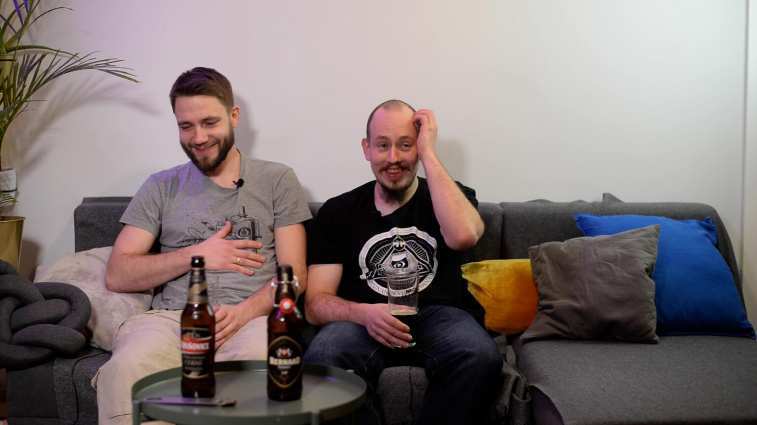 Grzegorz Stachurski (po lewej) i Łukasz Szymański (po prawej) siedzą na kanapie. Grzegorz trzyma się za brzuch, Łukasz łapie za głowę ze śmiechu, Przed nimi stoi krągły metalowy stolik, na którym stoja 2 butelki TMavego.