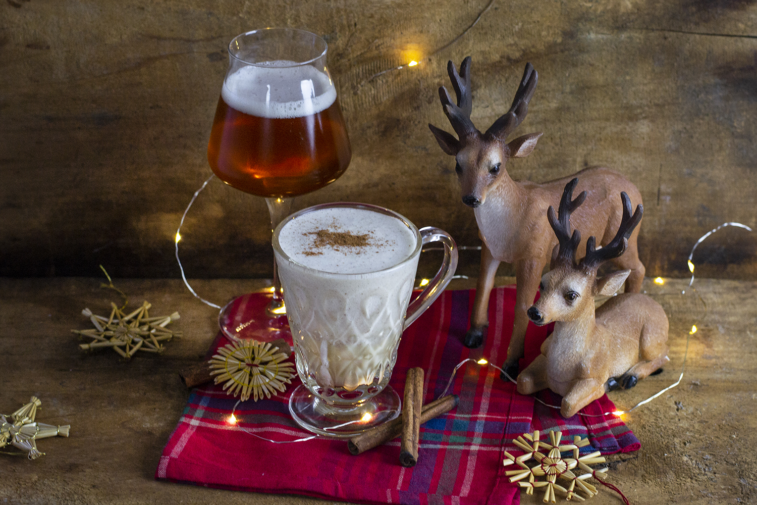 kompozycja świąteczna, kubek z egg nogiem przyozdobiony lampkami, obok renifery oraz szklanka piwa dyniowego. całość ozdobiona świątecznymi słomianymi ozdobami