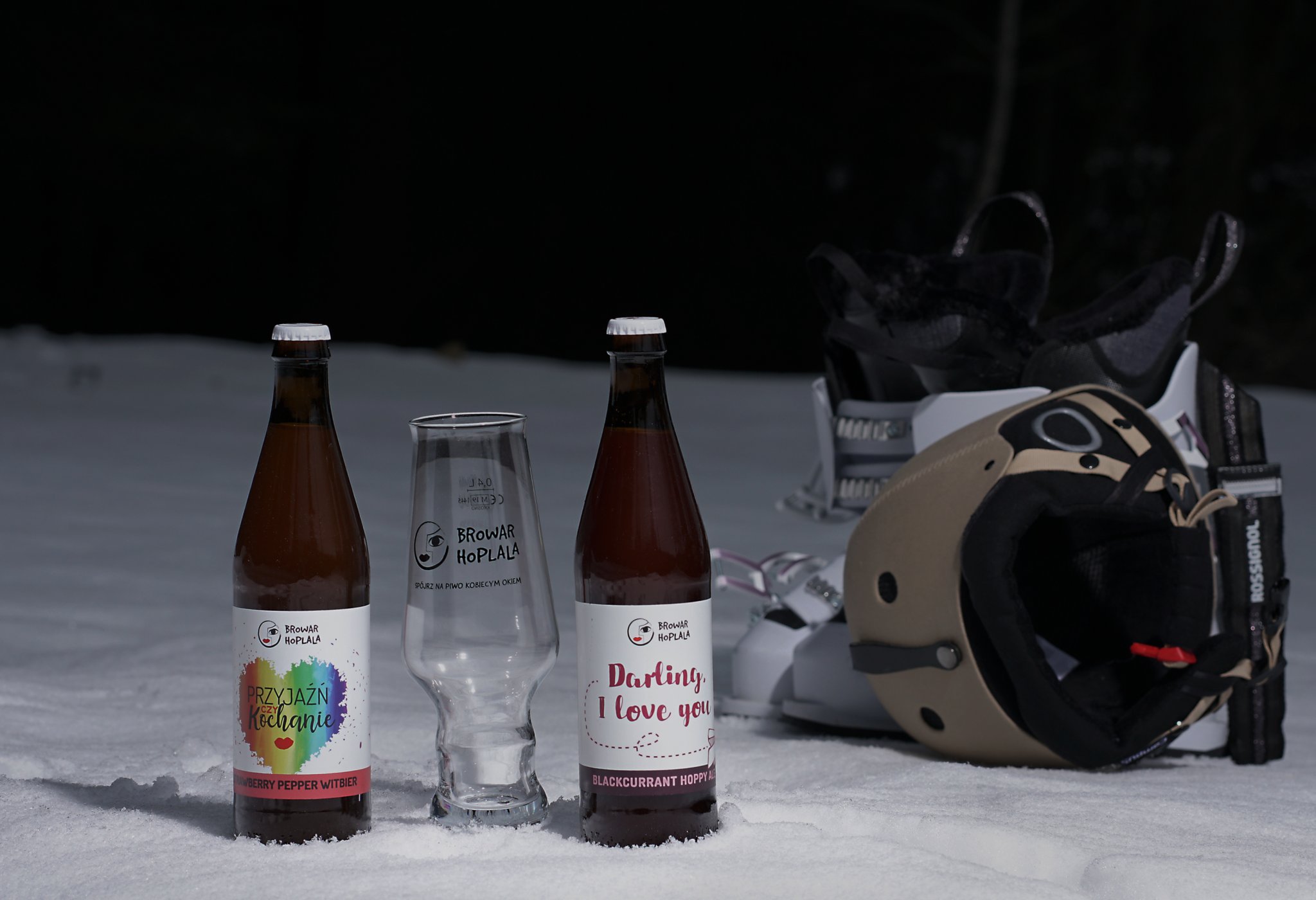 Dwie butelki piwa z hop lala - przyjaźń czy kochanie i Darling i Love you stojąće na śniegu w nocy. w tle buty narciarskie i kask narciarski